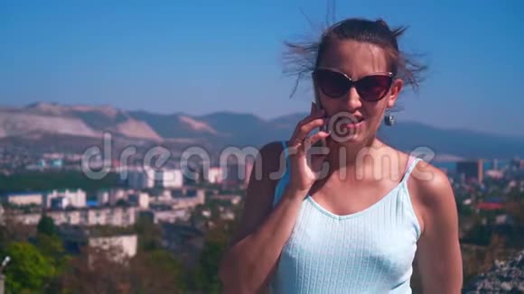 年轻迷人的女人戴着眼镜穿着蓝色的裙子站在城市的背景上在电话里说话视频的预览图