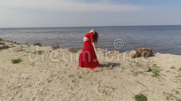 一位穿着红色长裙手舞足蹈的可爱年轻女子在蓝色海景附近的大石头上举手视频的预览图