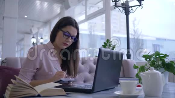 远程在线学习女学生在笔记本上写笔记用手提电脑坐在桌旁看书视频的预览图