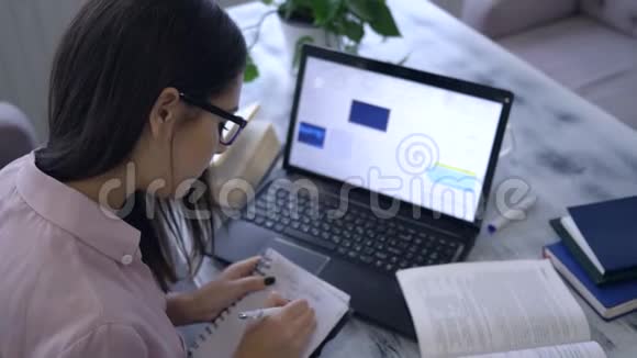 在线课程迷人的女孩在笔记本上写笔记看书坐在桌上用笔记本电脑视频的预览图