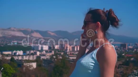 年轻迷人的女人戴着眼镜穿着蓝色的裙子站在城市的背景上在电话里说话视频的预览图