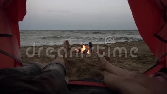轻松无忧无虑年轻人躺在沙滩上的红色帐篷里有海景和篝火的男女腿视频的预览图