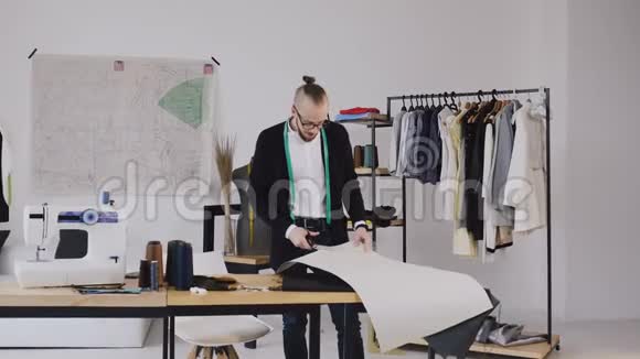 年轻的男设计师脖子上挂着胶带线站在服装制作工作室用剪刀剪成一个新的图案视频的预览图