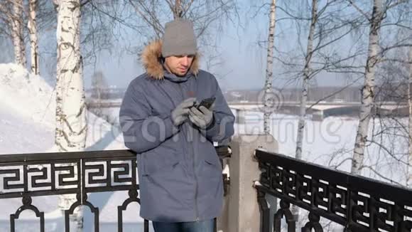 穿着蓝色夹克带着毛皮罩的人在冬季公园里用手戴手套擦拭手机屏幕视频的预览图