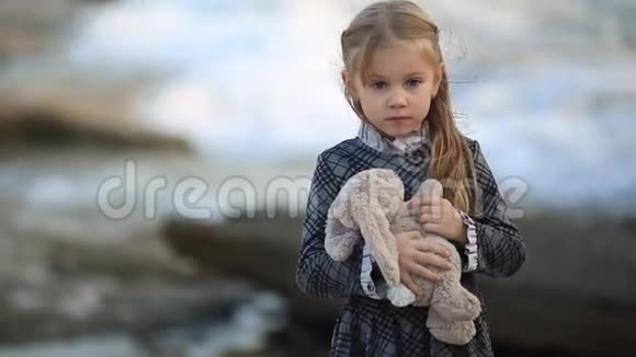 在一个刮风的日子里金发的小女孩站在海边女孩玩玩具兔子和软泰迪熊视频的预览图
