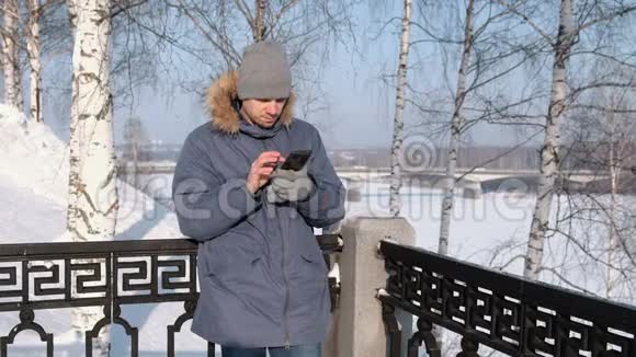 穿着蓝色羽绒服带着皮帽的人在冬季公园用手机上网视频的预览图