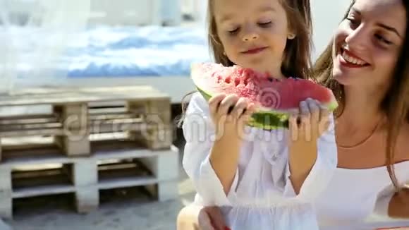 快乐开朗的小女孩穿着白色连衣裙和妈妈一起在海滩上吃西瓜玩得很开心视频的预览图