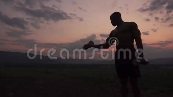 非洲裔美国人肌肉健美的身体建设者的剪影举着哑铃在粉红色日落的天空背景室外视频的预览图
