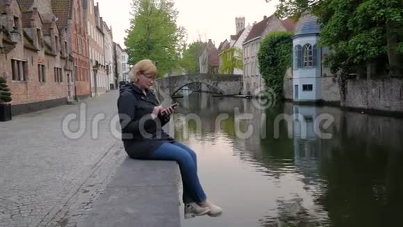黑色外套中的成熟女商人使用电话应用程序坐在河边频道视频的预览图