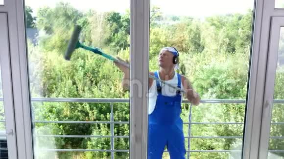 透过窗户观看英俊的男清洁工人穿着蓝色工作服带着耳机清洁洗涤视频的预览图