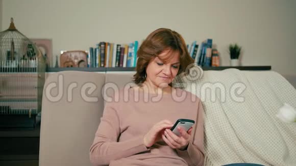 漂亮的成年女性在舒适的房间里坐在沙发上的智能手机上使用应用程序视频的预览图