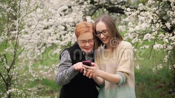 年长的母亲和她的成年女儿在盛开的花园里看照片视频的预览图