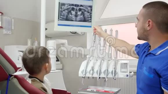 医学和医疗保健可爱的正畸医生与孩子在牙科扶手椅交谈并显示他的手放在监视器上视频的预览图