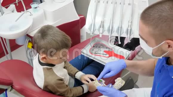 专业医生戴着医疗面具在模型下巴上向坐在牙科扶手椅上的小个子展示牙板视频的预览图