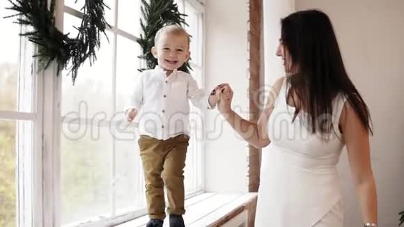 年轻微笑的母亲穿着白色连衣裙正牵着儿子手而他走在装饰着的窗台上视频的预览图