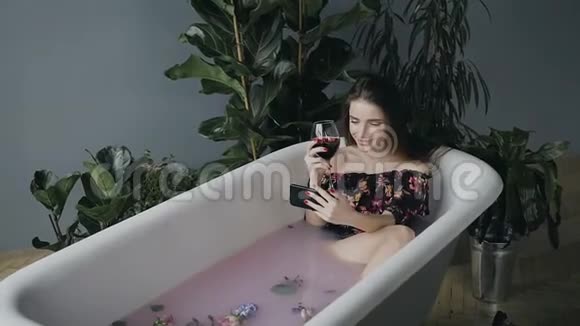 一个拿着一杯红酒的女孩坐在一个温暖的奶浴中用芳香的花蕾和电话放松视频的预览图