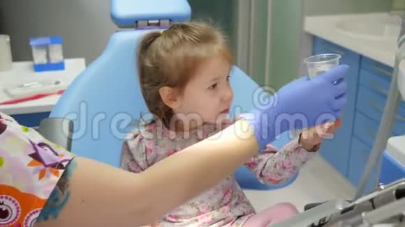 可爱的小女孩喝着塑料杯里的水坐在椅子上坐在橱柜里的女医生旁边视频的预览图