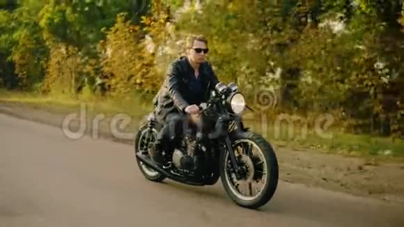 秋天的森林里阳光明媚的一天穿着太阳镜和皮夹克的时髦男人骑着酷酷的自行车在柏油路上视频的预览图