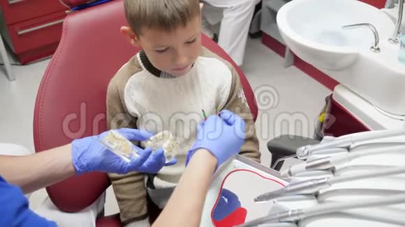 专业口腔医生为好奇的孩子在牙科扶手椅上展示一个模型下巴和牙板视频的预览图