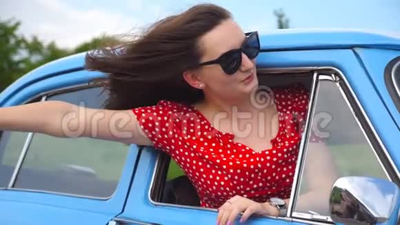 年轻女孩戴着太阳镜斜靠在老式车窗外享受旅行女人从复古汽车中脱颖而出视频的预览图
