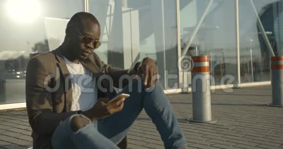 有魅力的现代非洲男人戴着太阳镜坐在路上靠在地上一边通过手机说话视频的预览图