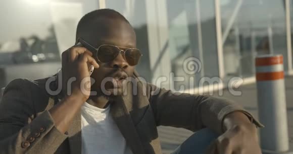 穿着时尚太阳镜的迷人的非洲男人坐在路上时情绪激动地交谈和做手势视频的预览图