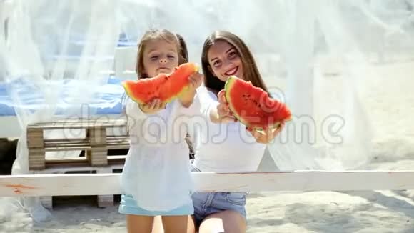快乐开朗的小女孩穿着白色连衣裙和妈妈一起在海滩上吃西瓜玩得很开心视频的预览图