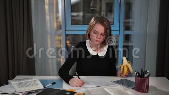 一个快乐的女学生用平板电脑吃香蕉在笔记本上写练习午休时间她做她视频的预览图