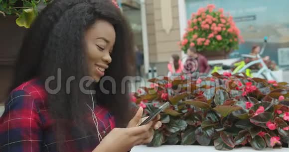 有趣的积极的非裔美国女孩带着漂亮的微笑听着耳机里的音乐用手机聊天视频的预览图