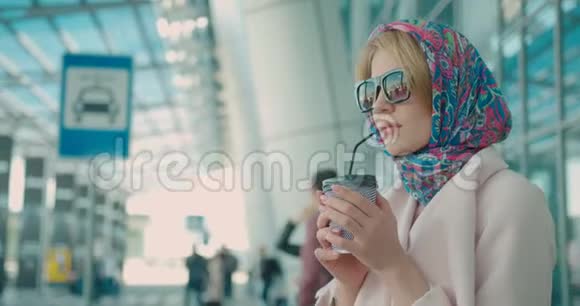 穿着米色外套戴着太阳镜戴着围巾的时髦年轻女性正从她的金发上穿过小管喝咖啡视频的预览图