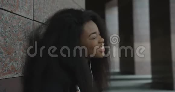 侧写一个情绪激动的非洲女孩正靠在大楼里一边用耳机欣赏音乐4k视频的预览图