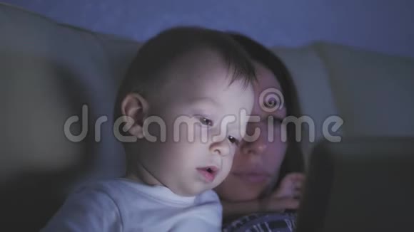 晚上妈妈和小儿子坐在家里的沙发上玩数字平板电脑视频的预览图