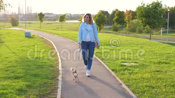 白种人女人带着他的小狗在绿色公园的夕阳下走出去视频的预览图
