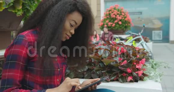 这位年轻迷人的非洲女孩在户外坐着一边通过平板电脑聊天浏览和发短信一边平静地散步视频的预览图