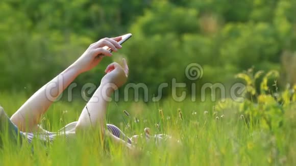 躺在绿色草坪上的快乐年轻女子在夕阳背景下的风景区使用智能手机视频的预览图