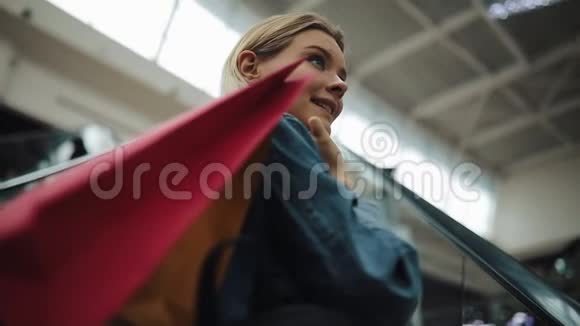 年轻的金发女郎穿着牛仔裤夹克微笑着走上购物中心的移动楼梯视频的预览图