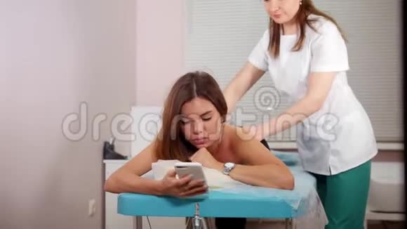 一位年轻女子接受放松的背部按摩坐在她的电话里视频的预览图