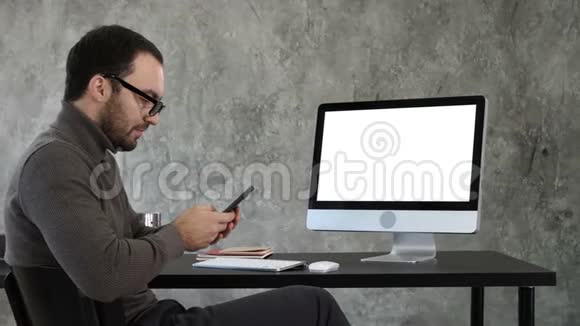 商人看着他的智能手机短信坐在电脑屏幕附近白色显示器视频的预览图