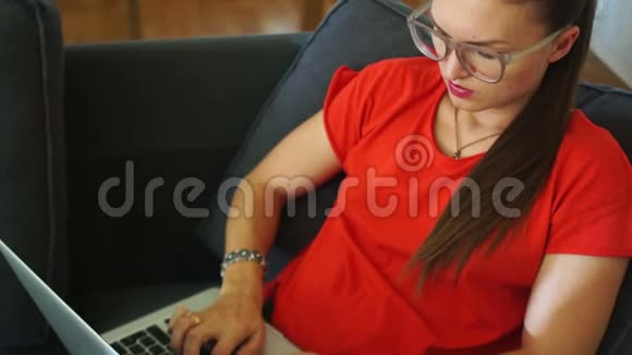 穿着红色连衣裙的黑发女人正坐在椅子上在笔记本电脑键盘上打字远程就业妇女视频的预览图