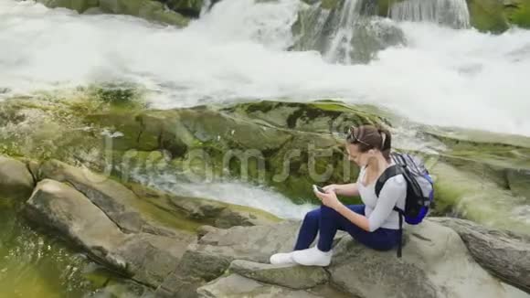 俯视图一位年轻女子坐在一块山河背景下的岩石上使用智能手机技术和视频的预览图