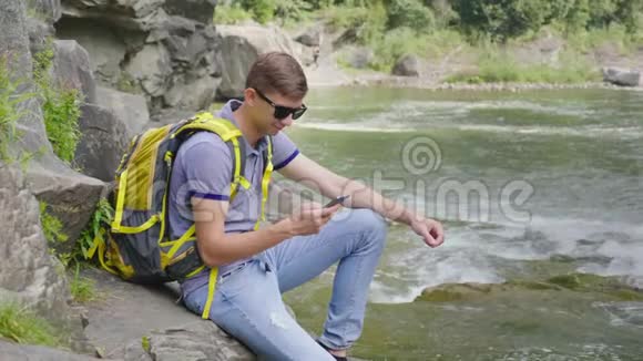 一个带着黄色背包的年轻人坐在一条山河附近使用智能手机旅游及科技概念视频的预览图