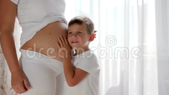 好奇的男孩有兴趣在白色窗帘的背景下倾听准妈妈的肚子视频的预览图