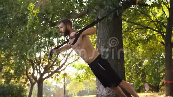 长胡子的年轻肌肉男在树附近的健身圈上做俯卧撑4K慢莫镜头耀斑效应视频的预览图
