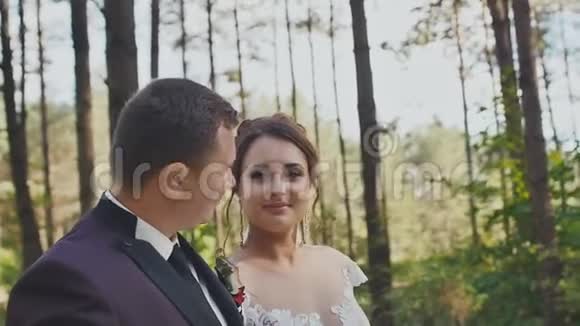 一位优雅的新郎和一位迷人的新娘带着一束明亮的花束沿着松树之间的森林小径散步快乐视频的预览图