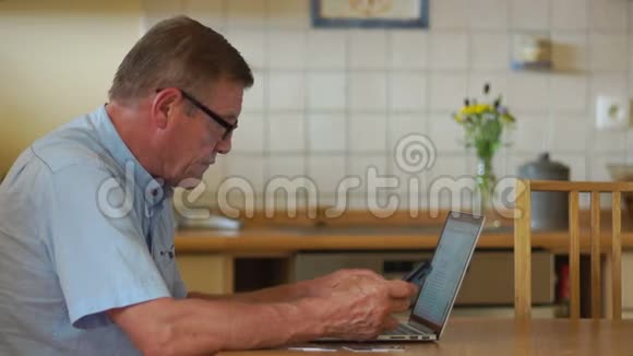 养老金领取者坐在厨房里用互联网和笔记本电脑浏览他的账户在桌子旁边是视频的预览图