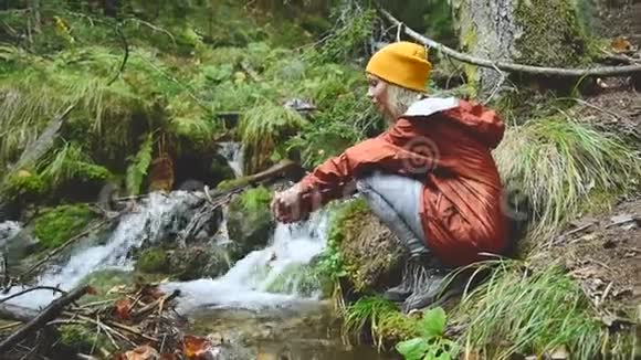 一个戴着帽子的漂亮女孩旅行者坐在针叶林中的一条山涧的岸边双手亮视频的预览图