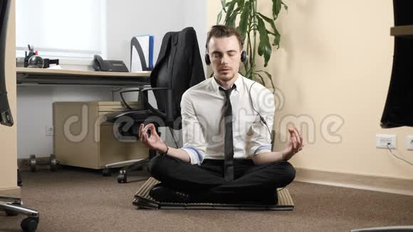 穿着西装戴着耳机坐在办公室中间的一个莲花姿势中冥想的年轻人视频的预览图