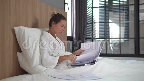 美丽的年轻女子在家坐在床上忙碌的少女穿着白色浴袍头发挽成发髻看上去视频的预览图