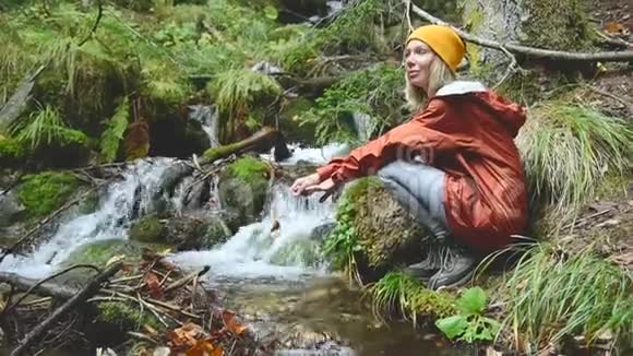 一个戴着帽子的漂亮女孩旅行者坐在针叶林中的一条山涧的岸边双手亮视频的预览图