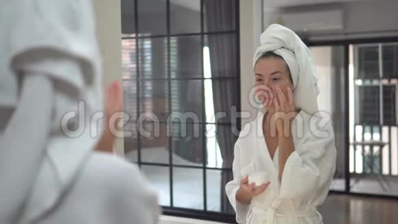漂亮漂亮的年轻女性站在浴室的镜子附近照顾她的皮肤在她的脸上涂上奶油视频的预览图